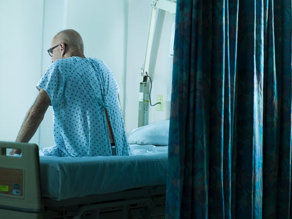 A man sits on the edge of a hospital bed, his back to the camera.