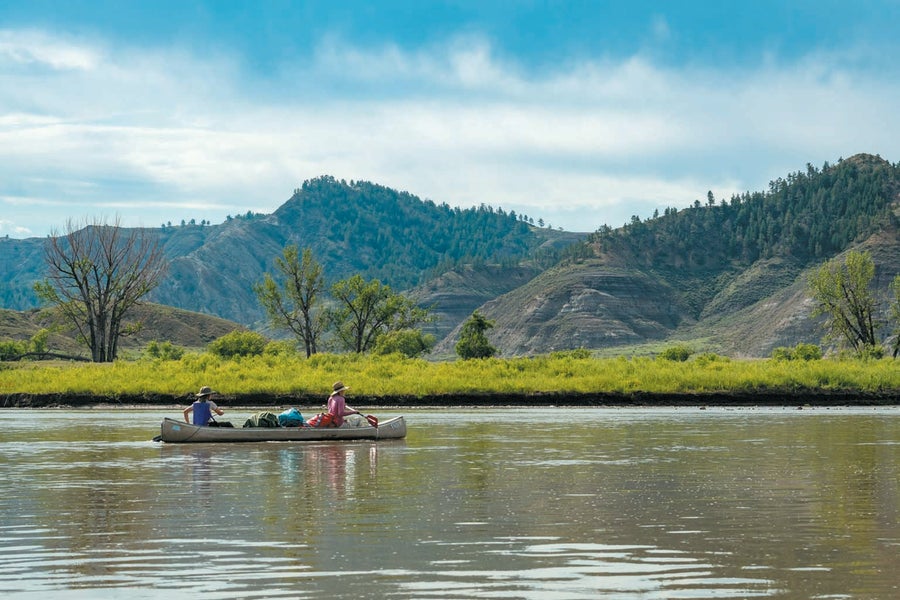A landscape with blue sky and clouds, mountains, vegetation and two people in a canoe in a body of water.