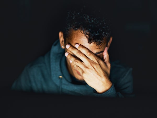 A hoodie-wearing young man, seen sitting in the dark, holds his hand over his face, exhausted