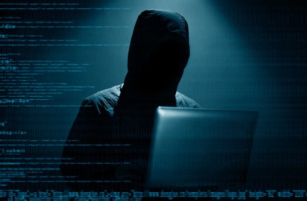 A hooded figure sits behind a computer with code displayed around him.