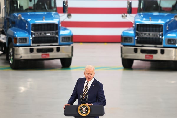 President Joe Biden speaking at a truck factory with two blue trucks and the US flag as background.