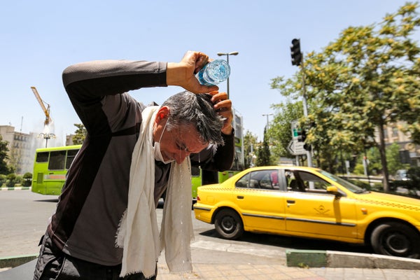 A man wearing a white scarf stands in front of a yellow car pouring water on his head from a bottle to cool off