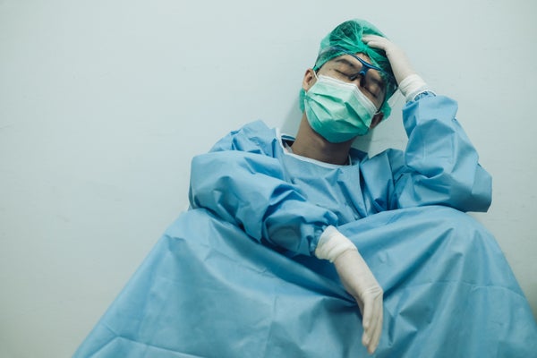 A male doctor in scrubs and a mask slumps against a wall, exhausted.ll