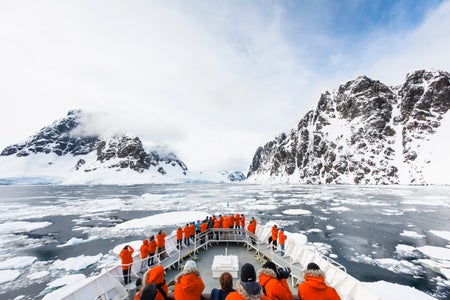 Passengers in bright orange coats on a ship's bow overlook an icy ocean