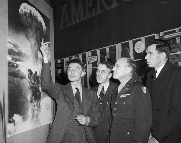 A black-and-white photograph of four men looking at a picture of an atomic bomb cloud.