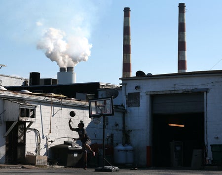 A person shoots basketball behind a dilapidated building. In the background, emissions spew from a coal-fired power plant.