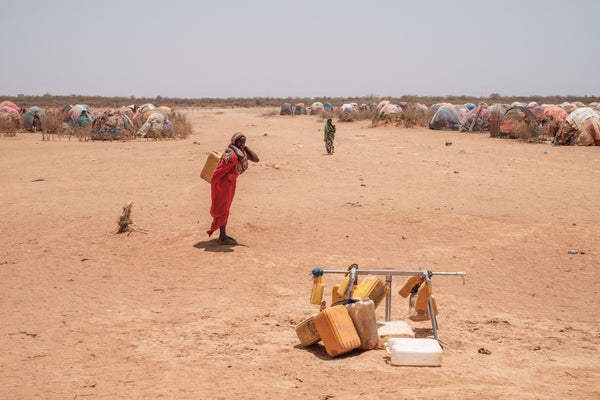 A woman stands next to out of service water taps on sand in Somalian camp.
