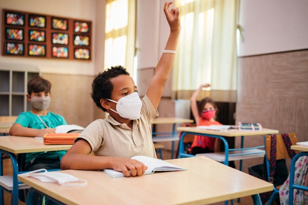 African-American schoolboy with protective mask is sitting at a desk in the classroom with raised hand.
