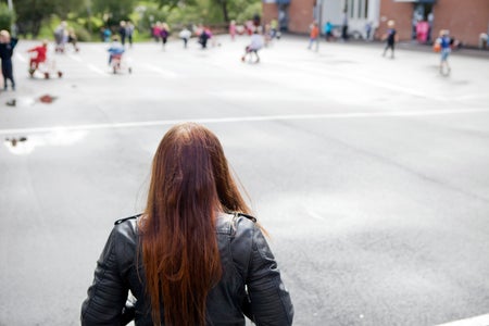A girl in a leather jacket looks out forlornly on a school yard.