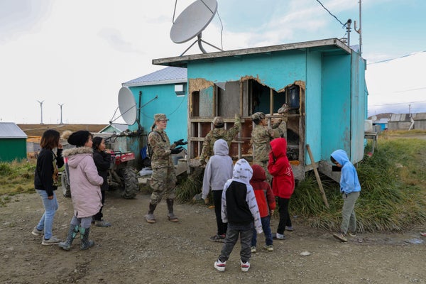 Soldiers and gather around the exterior of a damaged light blue building