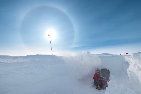 Person clears snow using snow blower from the entrance to a drill and science trench in Greenland.
