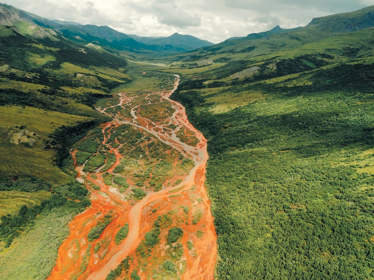  An aerial view of the orange-colored Copper River winding through a valley in Alaska, with green mountains rising on either side.