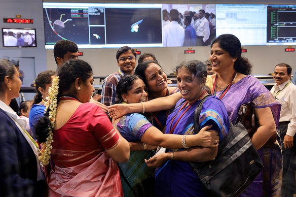 Indian staff from the Indian Space Research Organisation are seen celebrating in a command center. A group of five staff members wearing saris are centered at the front of the image's frame