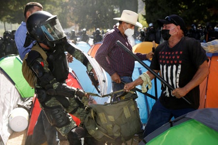 A farmer and a police officer in riot gear wrestle over bag