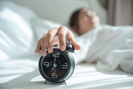 Female lying on bed reaches towards an alarm clock in foreground trying to turn it off