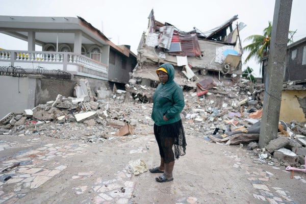 A sad woman stands in front of a collapsed house.