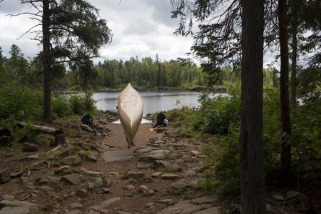 A person obscured by the canoe they are carrying stands close to a rocky shore in a heavily wooded area.