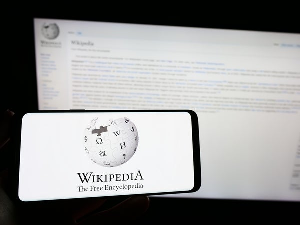 ··· Person holding smartphone with logo of online encyclopedia Wikipedia on screen in front of website. Focus on phone display.