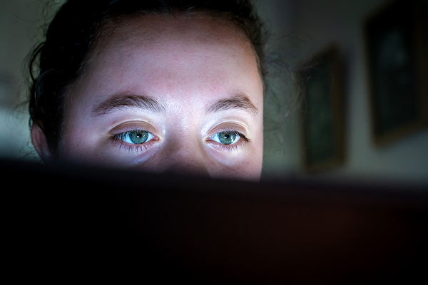 A young woman focuses on a computer screen that lights the top half of her face, with the bottom half in shadow.