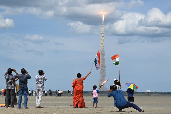 A rocket carrying Chandrayaan-3 lifts off from the spaceport in Sriharikota, in the foreground groups of adults and children watch, take photos, and wave Indian flags