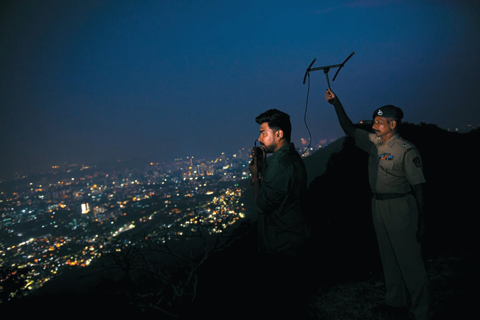 Two biologists are shown on an overlook of Mumbai, holding an antenna to track a radiocollared leopard.