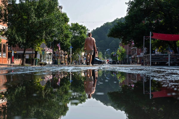 Man in a beige shirt and shorts walking down a flooded street - scene reflecting in water