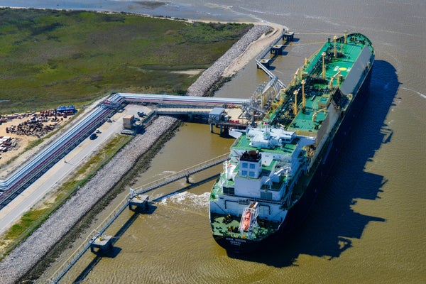 Aerial of LNG carrier ship sits docked with pipes connected at side.