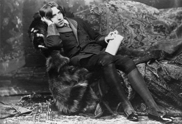 A man in Victorian clothing lounges on a fur-draped couch, resting his head on his right hand.