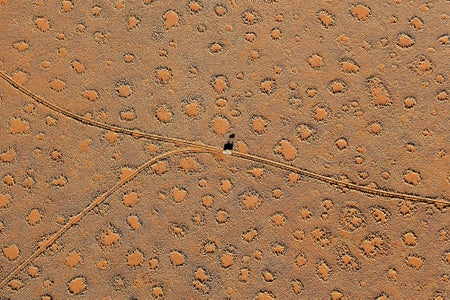 A vehicle photographed from a hot-air balloon driving on a road surrounded by fairy circles in the sandy landscape of western Namibia near low-lying mountain ranges