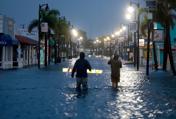 Two people wade through knee-deep water in the middle of a flooded commercial street