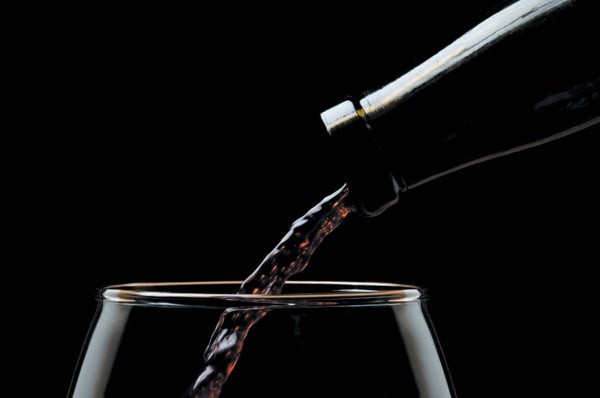 Studio photograph showing close-up of the top of a bottle of wine, with red wine pouring into a glass, with dramatic lighting.