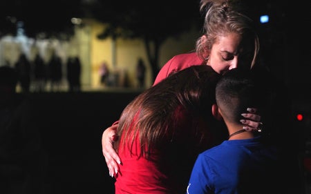 Mother in red shirt hugs her two children in red and blue shirts in front of school