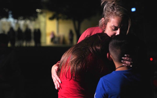 Mother in red shirt hugs her two children in red and blue shirts in front of school