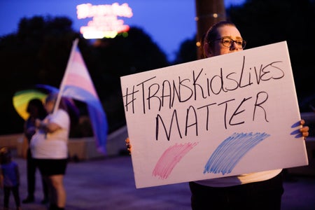 In the foreground, a transgender rights advocate holds a sign that reads, "#TRANSKIDSLIVESMATTER." In the background advocates can be seen holding a transgender pride flag and rainbow umbrella