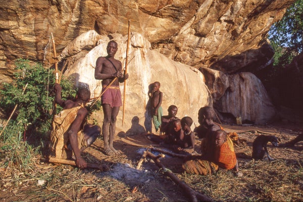 Men, women and children of The Hadza tribe gathered in front of rocks.