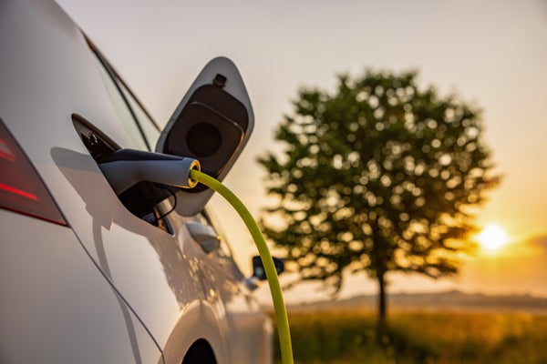Charging a white electric car on rural field with a tree background during sunset.