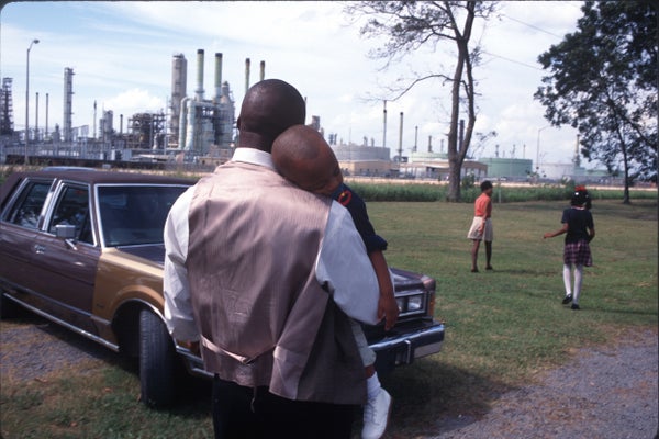 A black man holds a sleeping child as a woman and girl walk in the background, with a refinery looming nearby.