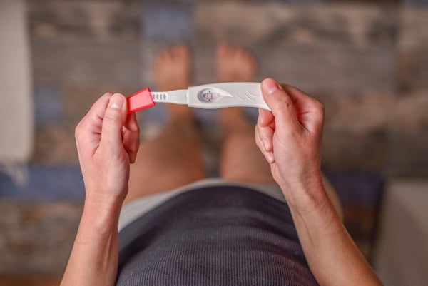 A standing woman looks down at a pregnancy test in her hands.