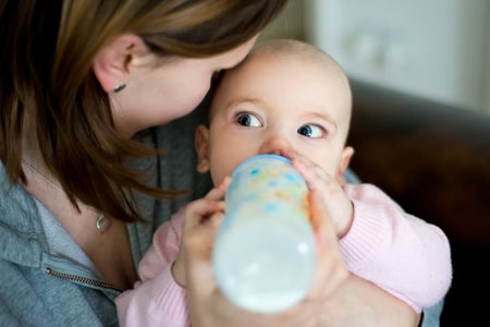 Baby girl with blue eyes and a pink sweater drinking milk from the bottle looking at her mum who is holding her and looking at her