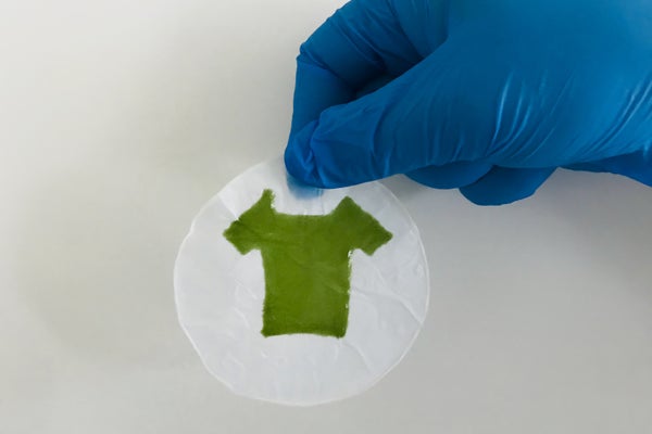 A gloved hand holds a round piece of material with a green, 3-D-printed T-shirt shape on it.