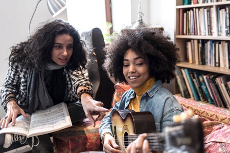 Young woman learning playing a guitar with teacher in cozy living room.