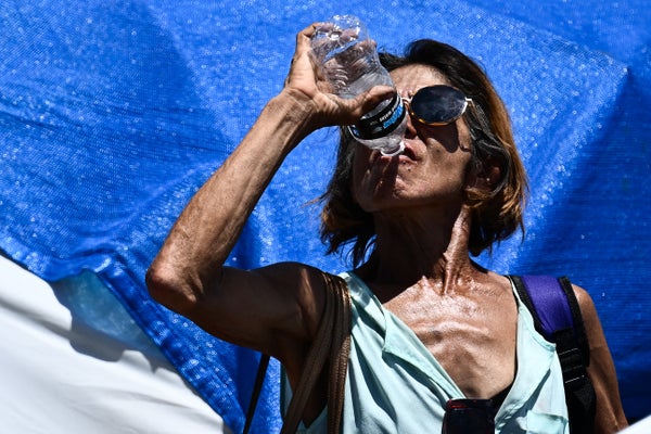 A person wearing sunglasses sweats as they drink from a plastic water bottle lifted high above their head, standing in bright sunlight in front of a blue tarp in the background