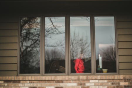 Window view of young girl in a bright colored shirt whose face is obscured by the reflection in the window of trees and sky.