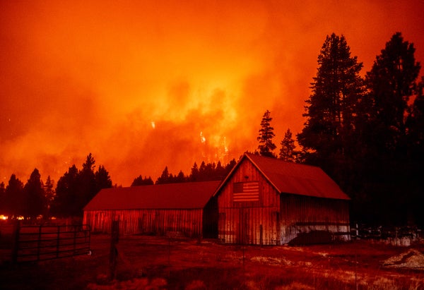 Flames rage behind a barn with an American Flag hanging on the wall