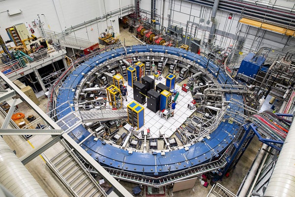 The Muon g-2 ring, viewed from above, sits in a large room, surrounded by various equipment. Two unidentified individuals stand in the middle and interact with equipment