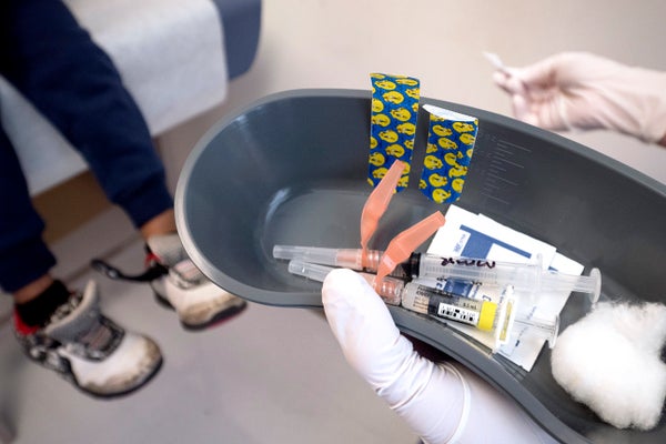 White gloves of a nurse holding vaccination items in a grey tray with feet of child in the background.