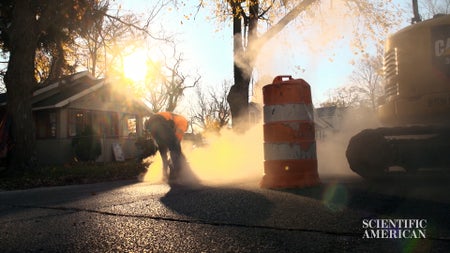 The sunlight pours through a cloud of dust created by a worker using a saw to cut through pavement.