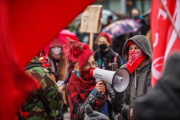 A woman with bright red hair shouts into a bullhorn, surrounded by other women and red banners.