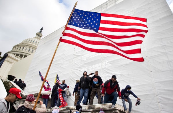 Men yelling below an American flag in front of the US Capitol building.