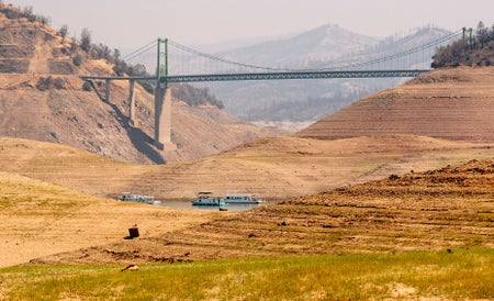 Boats sit at bottom of depleted river with bridge seen in distance and little water in sight.